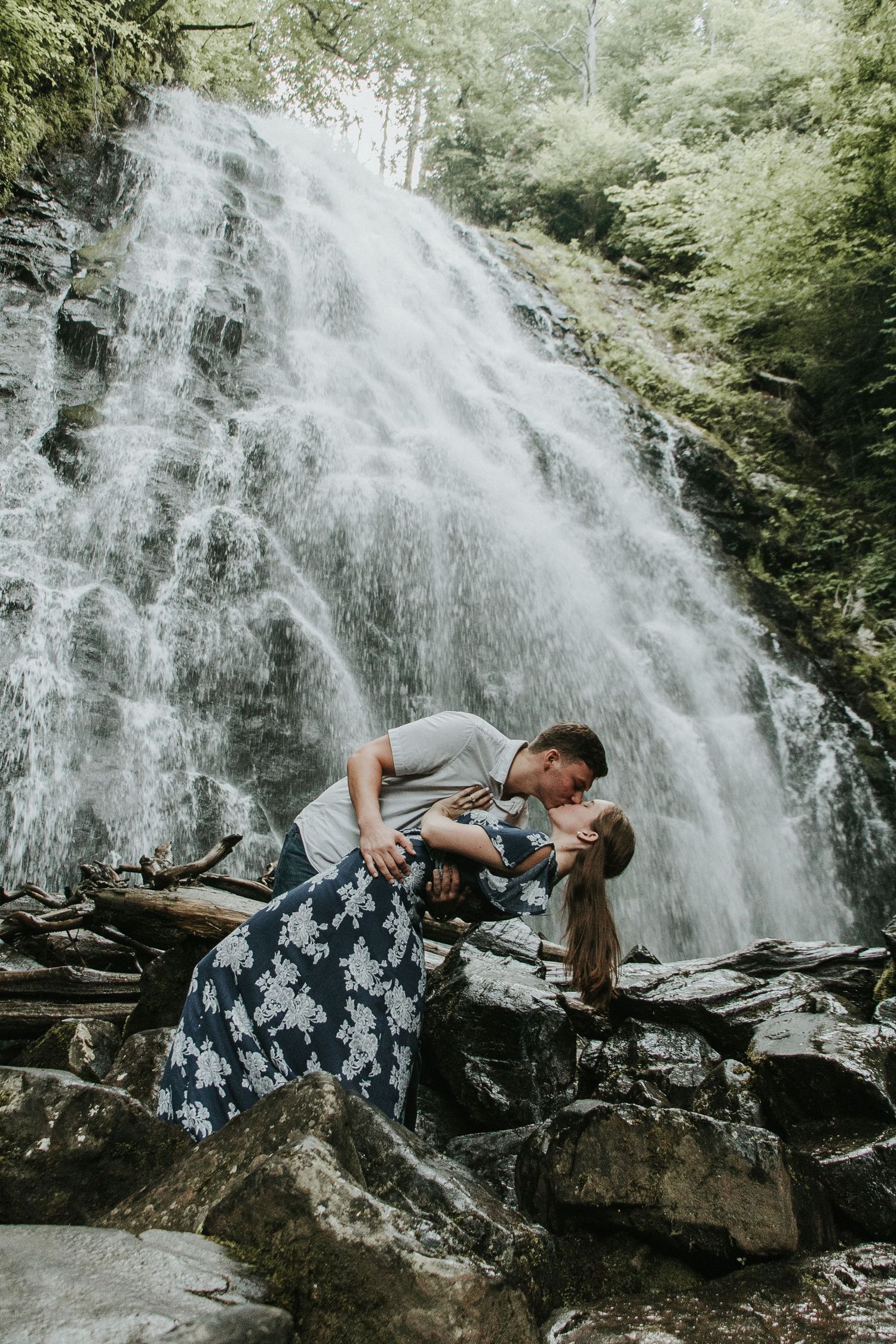 Romantic couple embracing in front of majestic waterfall on rocky terrain.