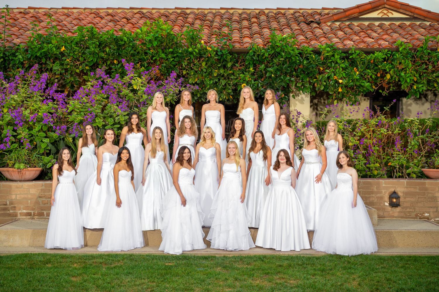 Large group in white dresses posed on garden steps with flowering purple wall behind them.