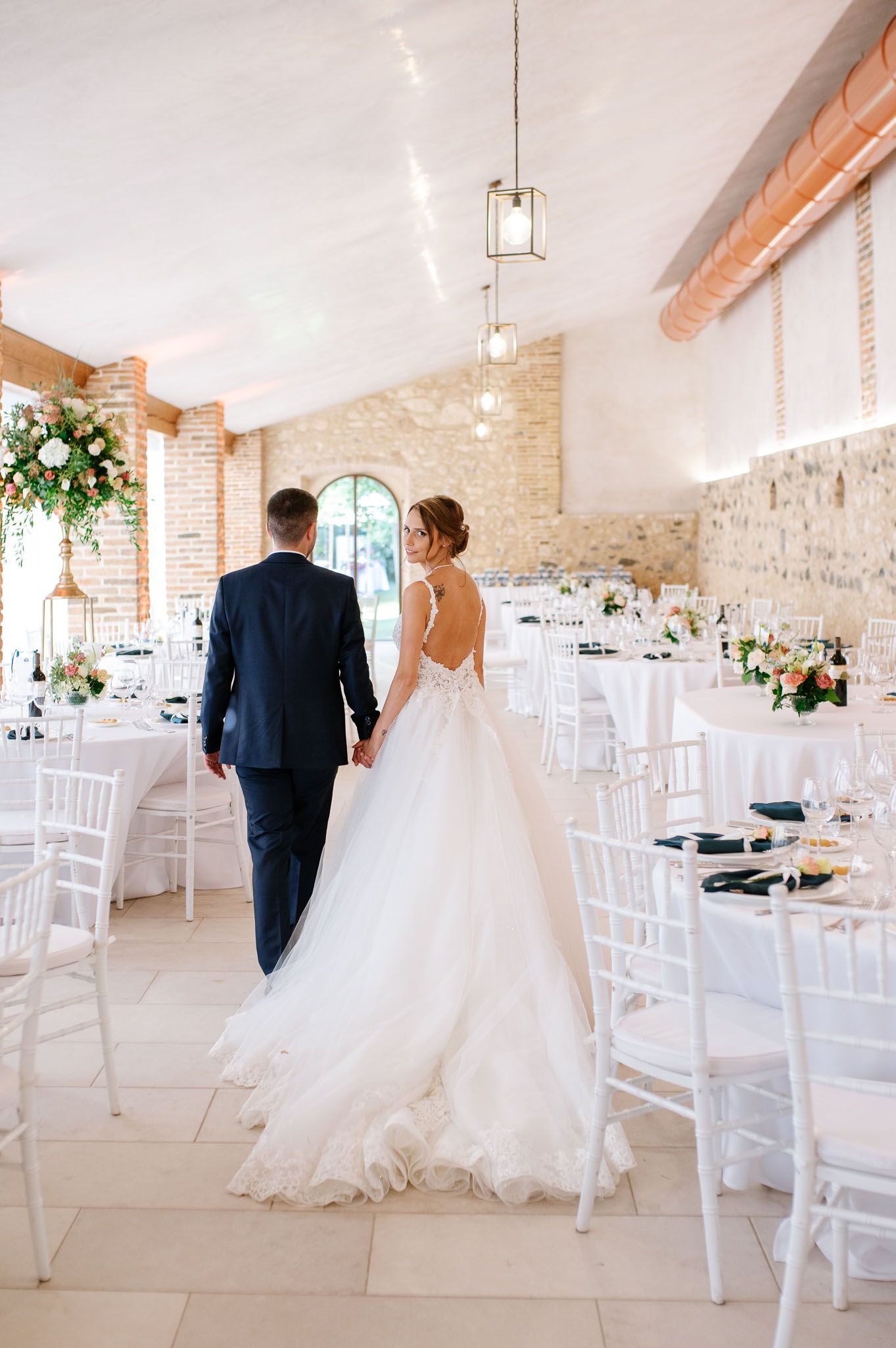 Newlyweds walk through elegant reception venue with exposed brick walls and white decor.