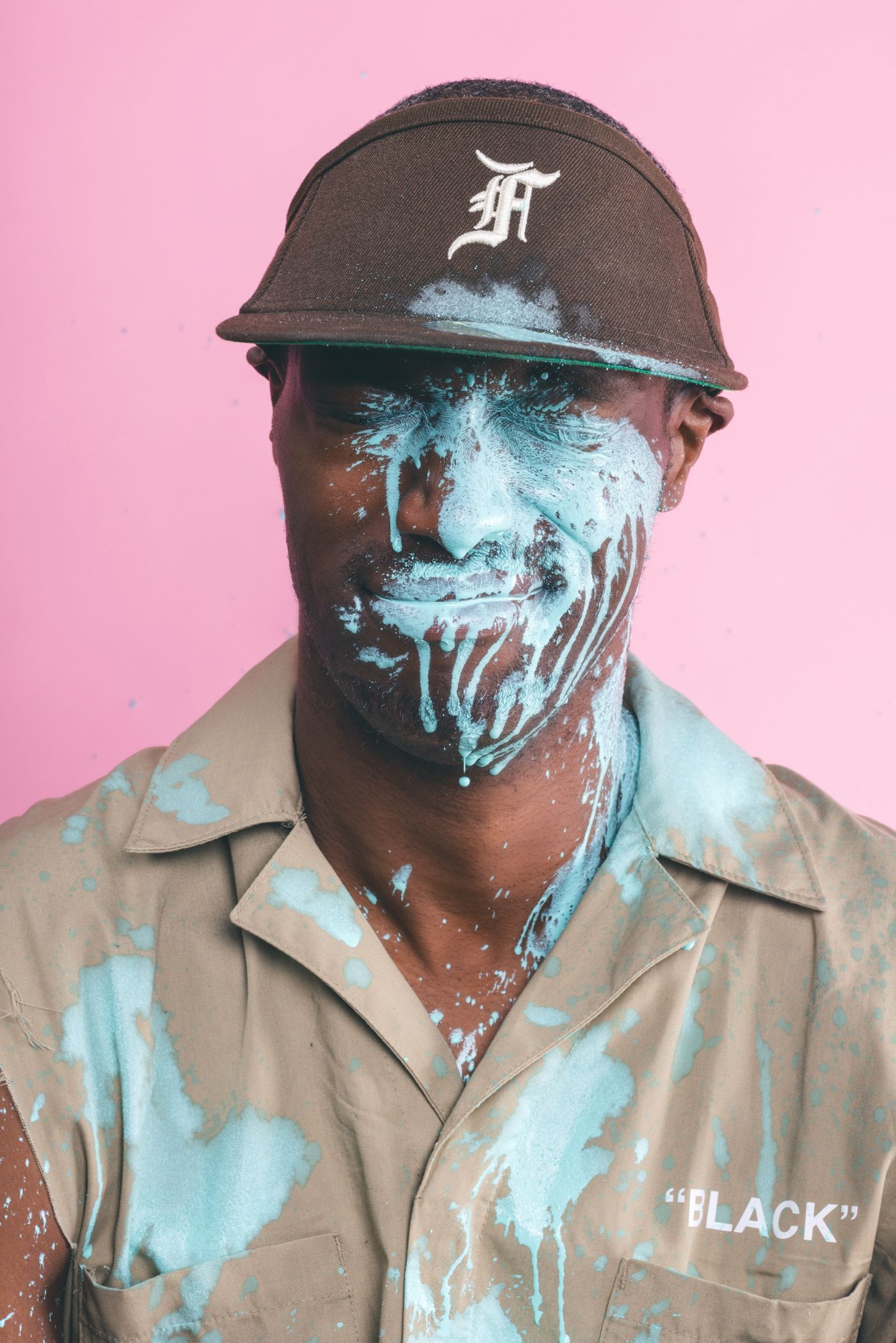 Person in baseball cap and tan jacket covered in bright blue paint against pink background.