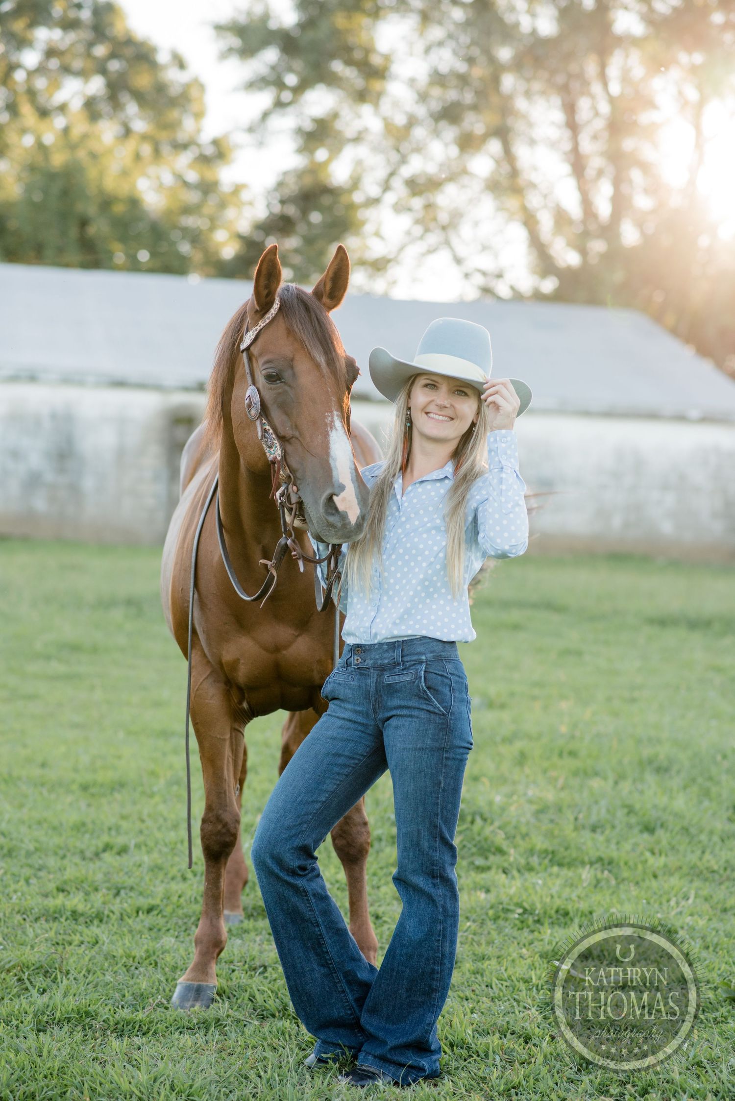 A person in western attire poses with a brown horse during golden hour in a grassy field.