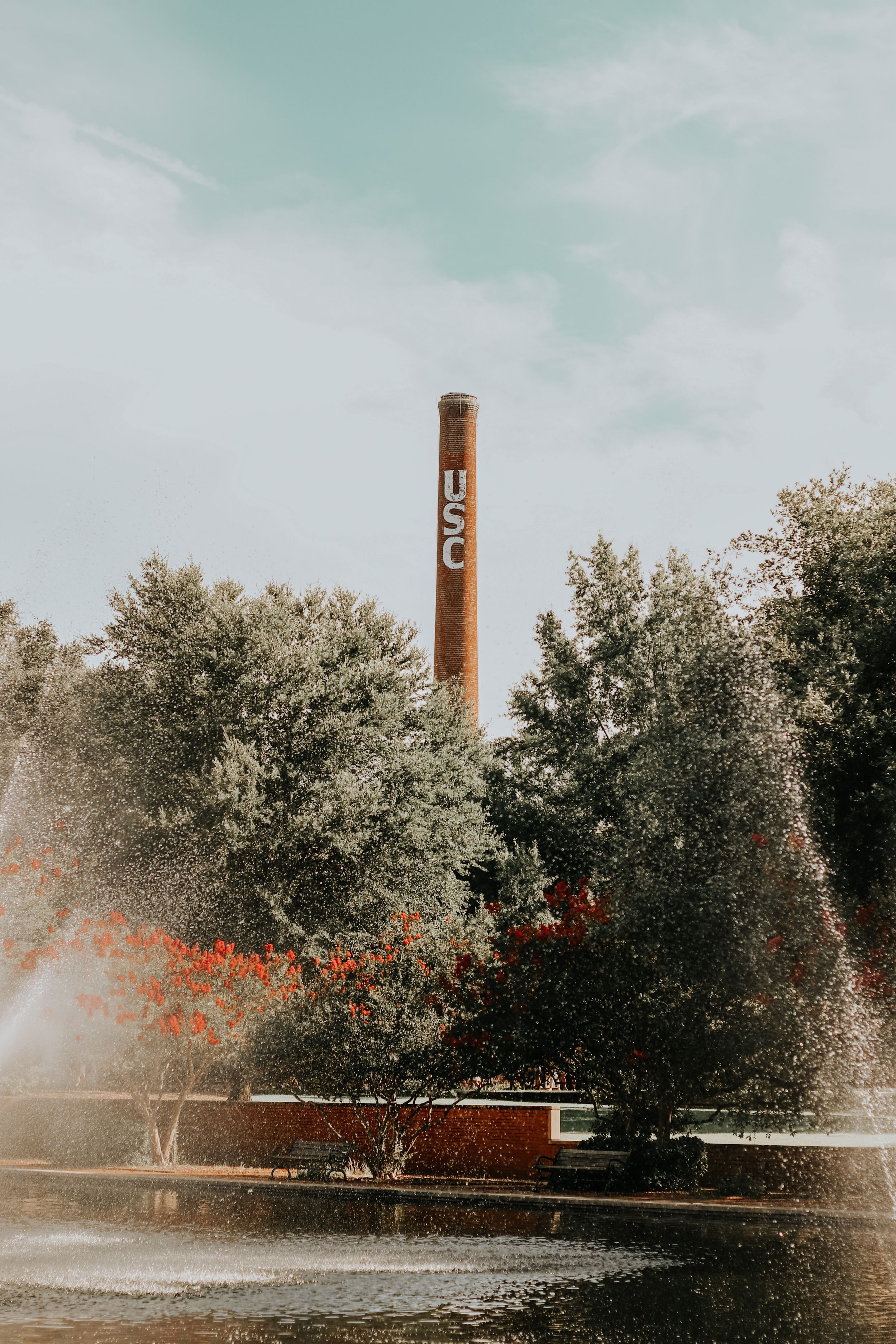 Tall brick industrial smokestack rising above trees against a light blue sky.