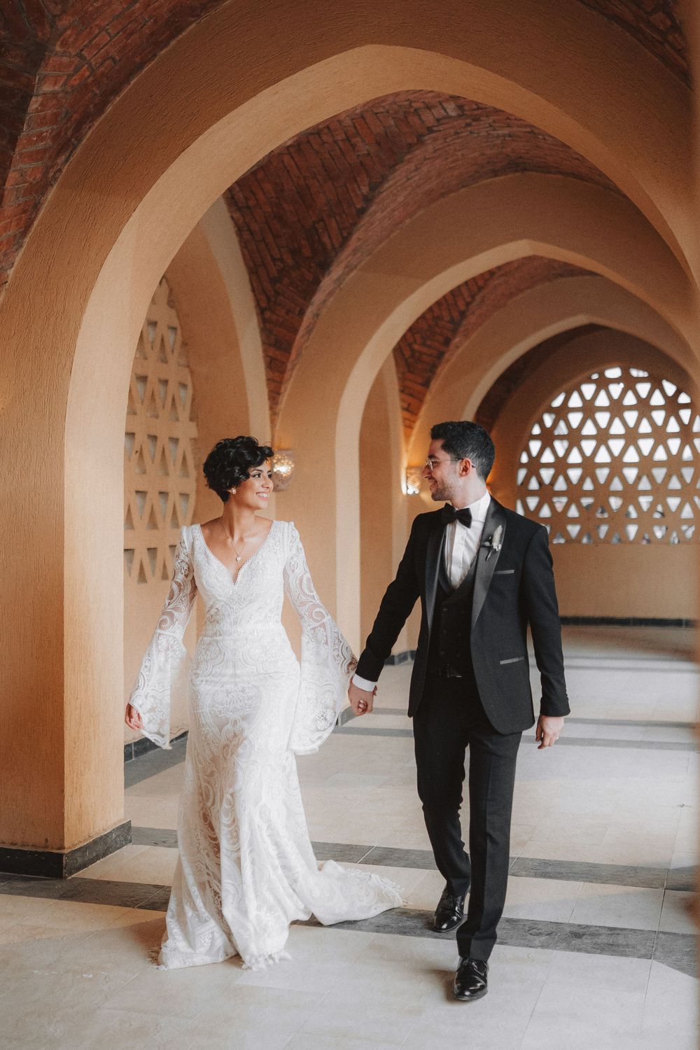 Couple in formal attire walking and posing playfully under arched hallway with decorative lattice windows.