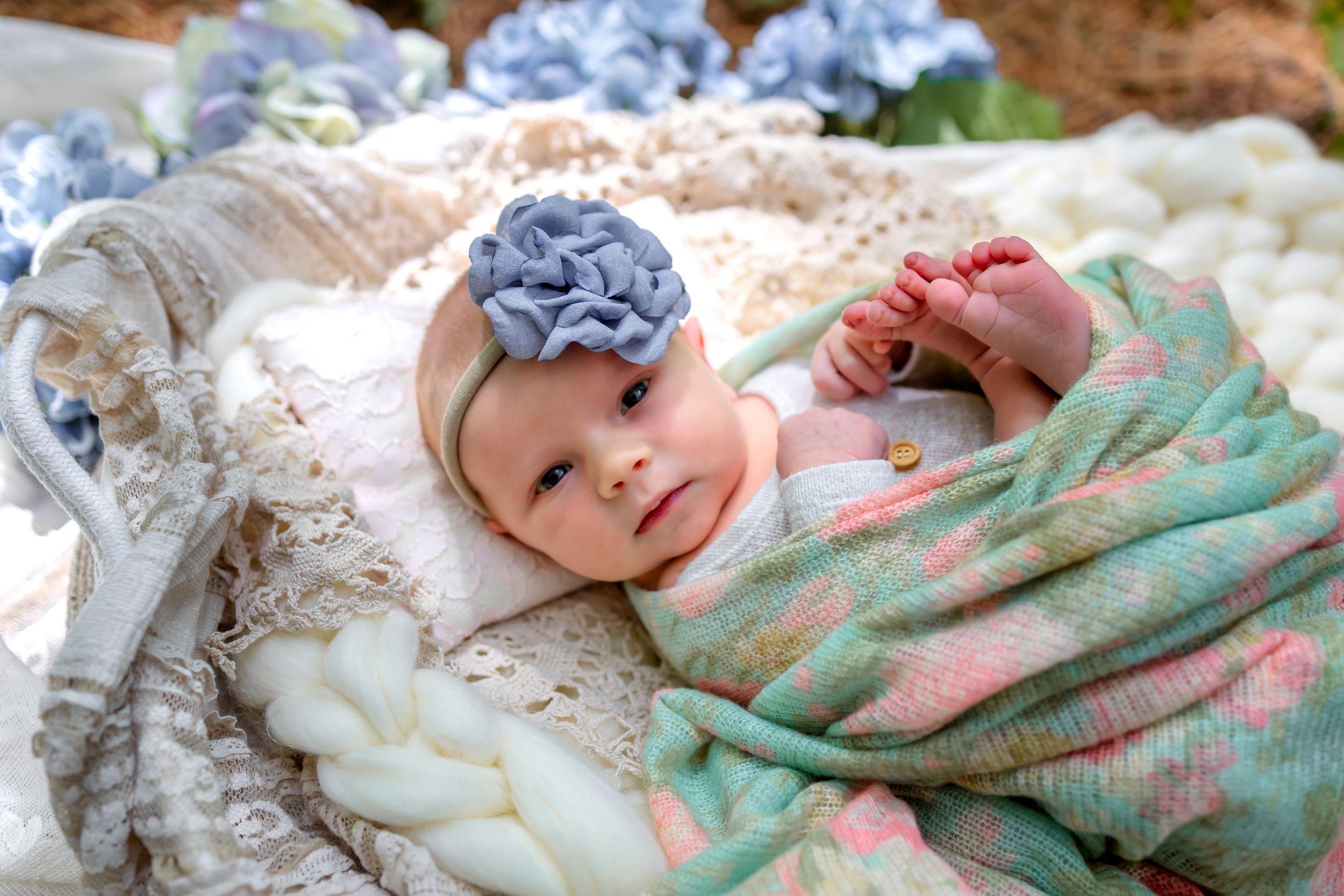 A baby in a floral headband lies on a soft blanket with a dreamy expression.