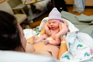 A newborn baby wearing a pink hat rests on their parent's chest in a hospital room.