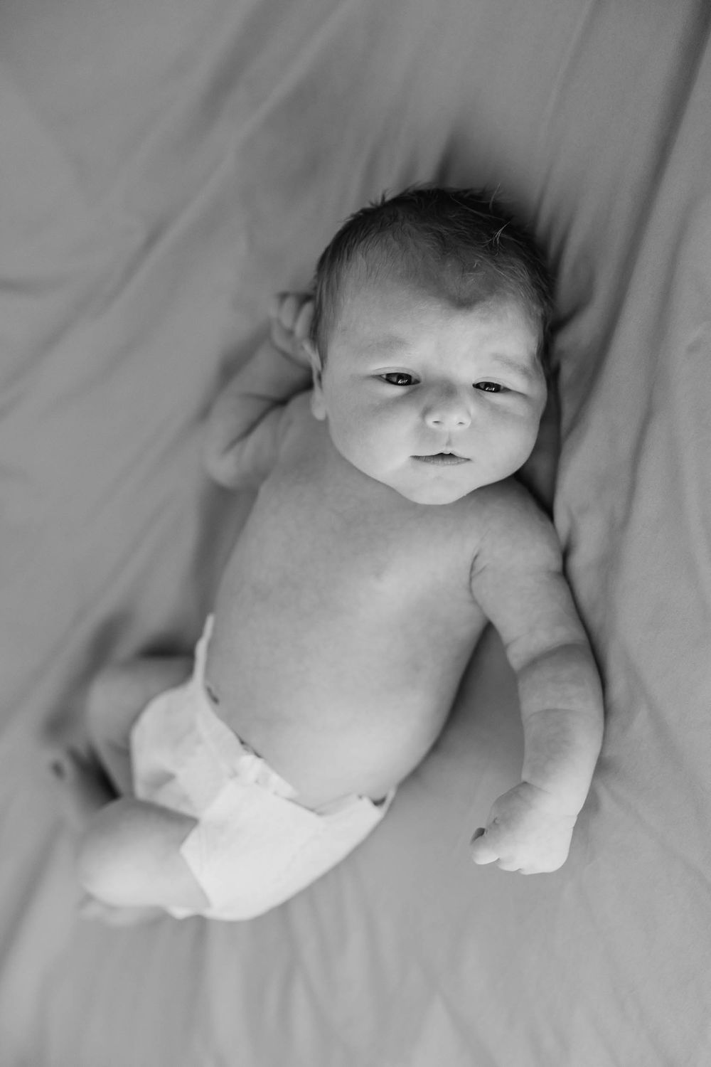A newborn baby wearing a diaper lies on soft white fabric in a black and white photograph.