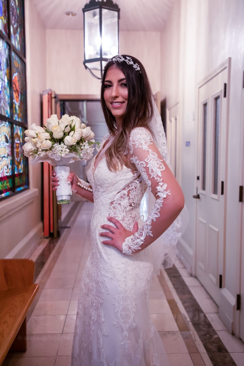A bride wearing a lace long sleeve wedding dress poses with bouquet in church hallway.