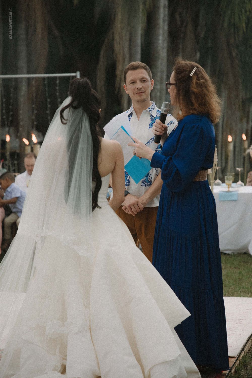 An outdoor wedding ceremony with couple and officiant in blue dress under palm trees.