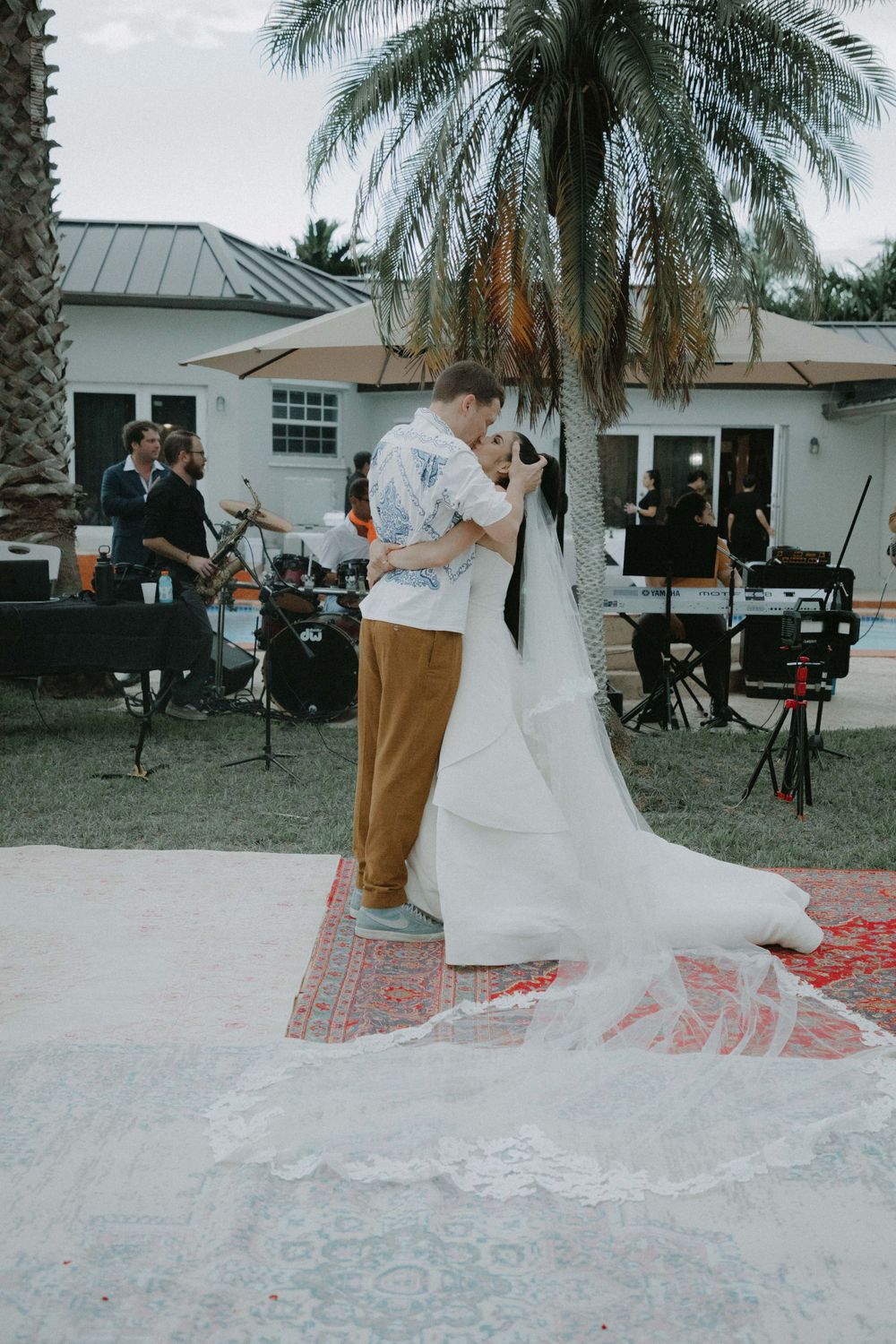 Couple dancing at an outdoor wedding reception under a palm tree with live band performing in the background.