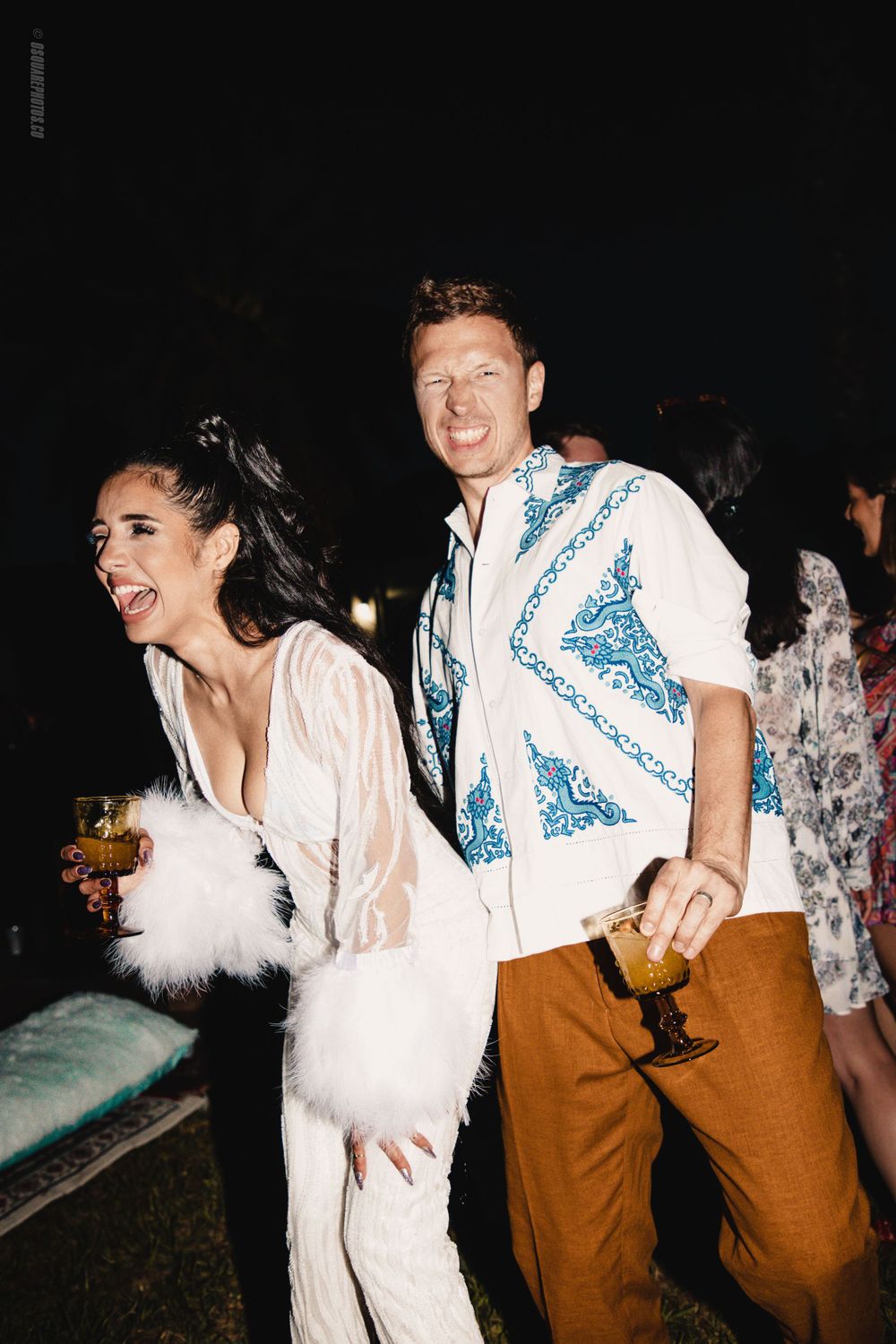 A couple in festive attire laughing and dancing at a nighttime party, with her in white and him in a blue-detailed shirt.