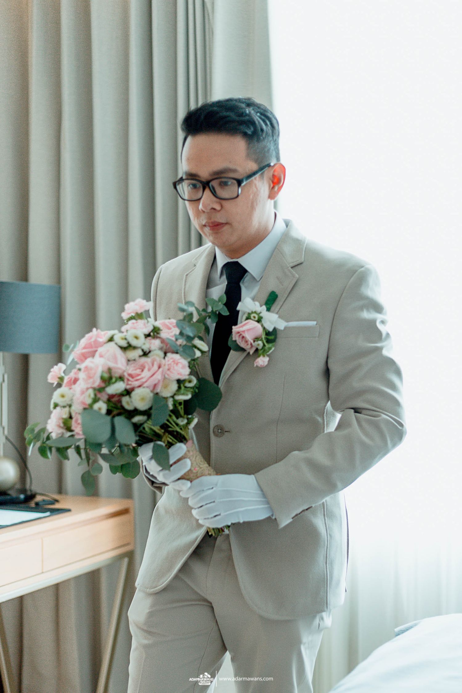 A groom in a light gray suit and glasses holds a pink and white floral bouquet.