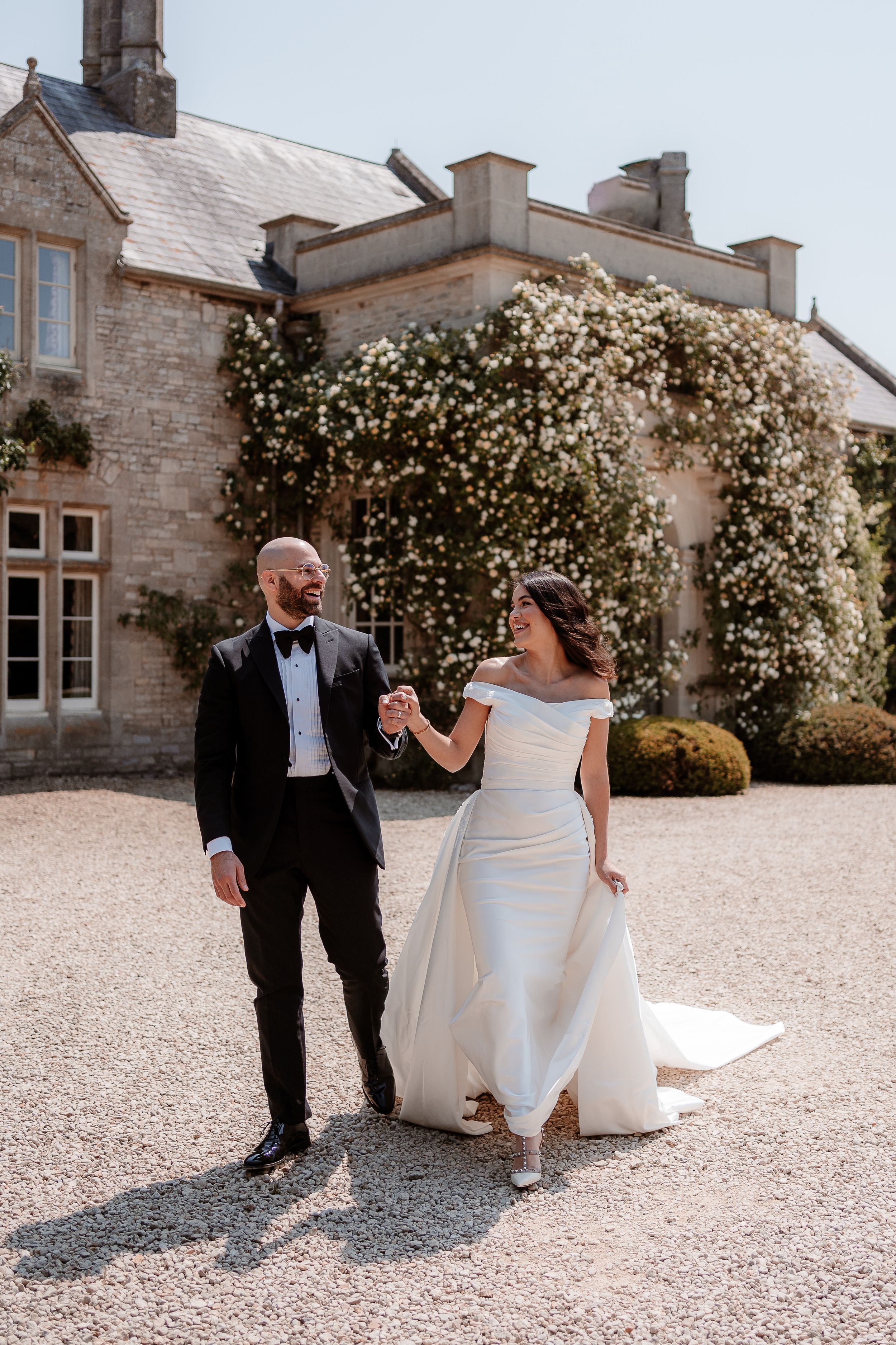 A couple laugh together, walking hand in hand by the rose covered entrance to Euridge Manor, Cotswolds wedding venue
