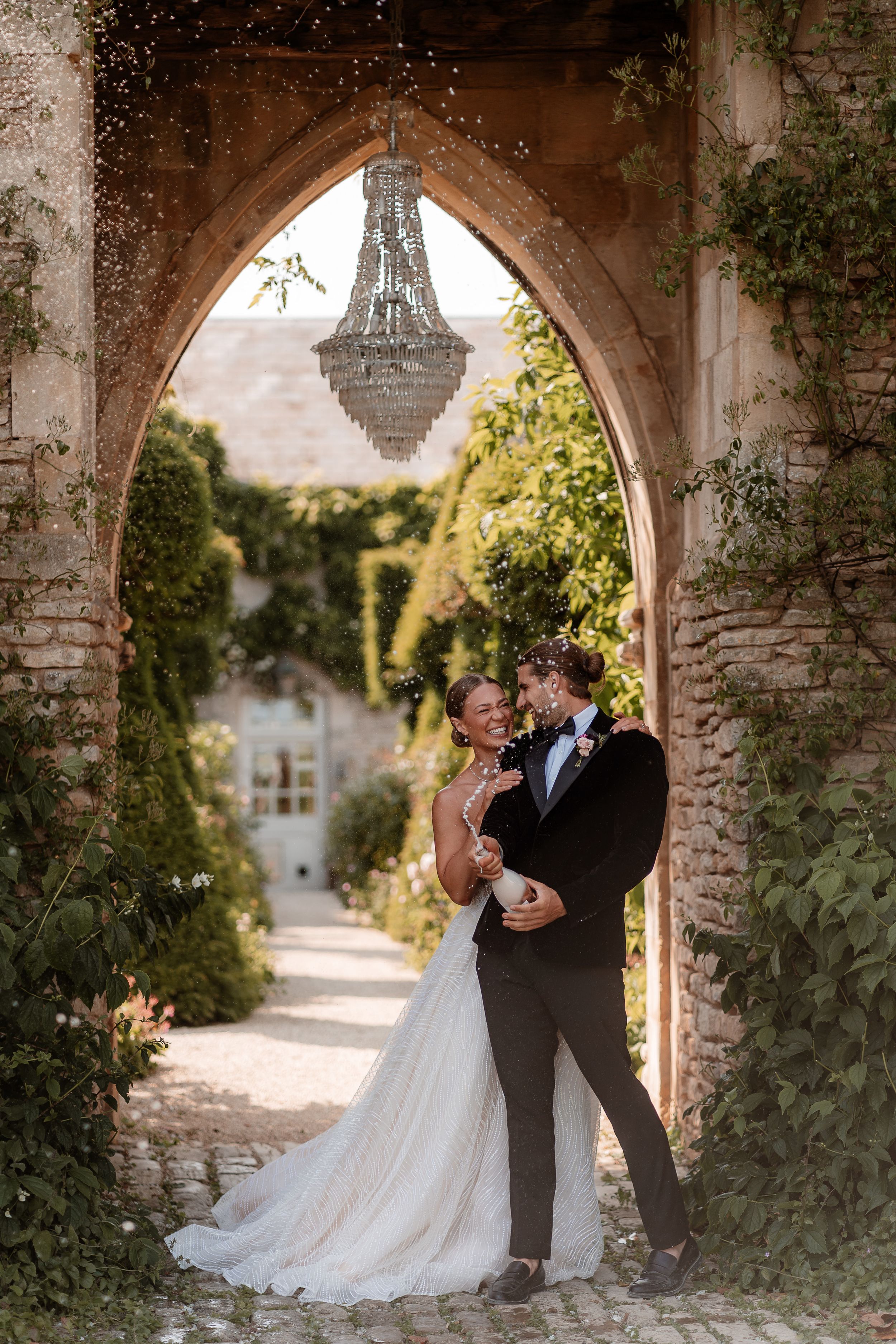A bride and groom spray a bottle of champagne together under the chandelier and castle arch at Euridge Manor.