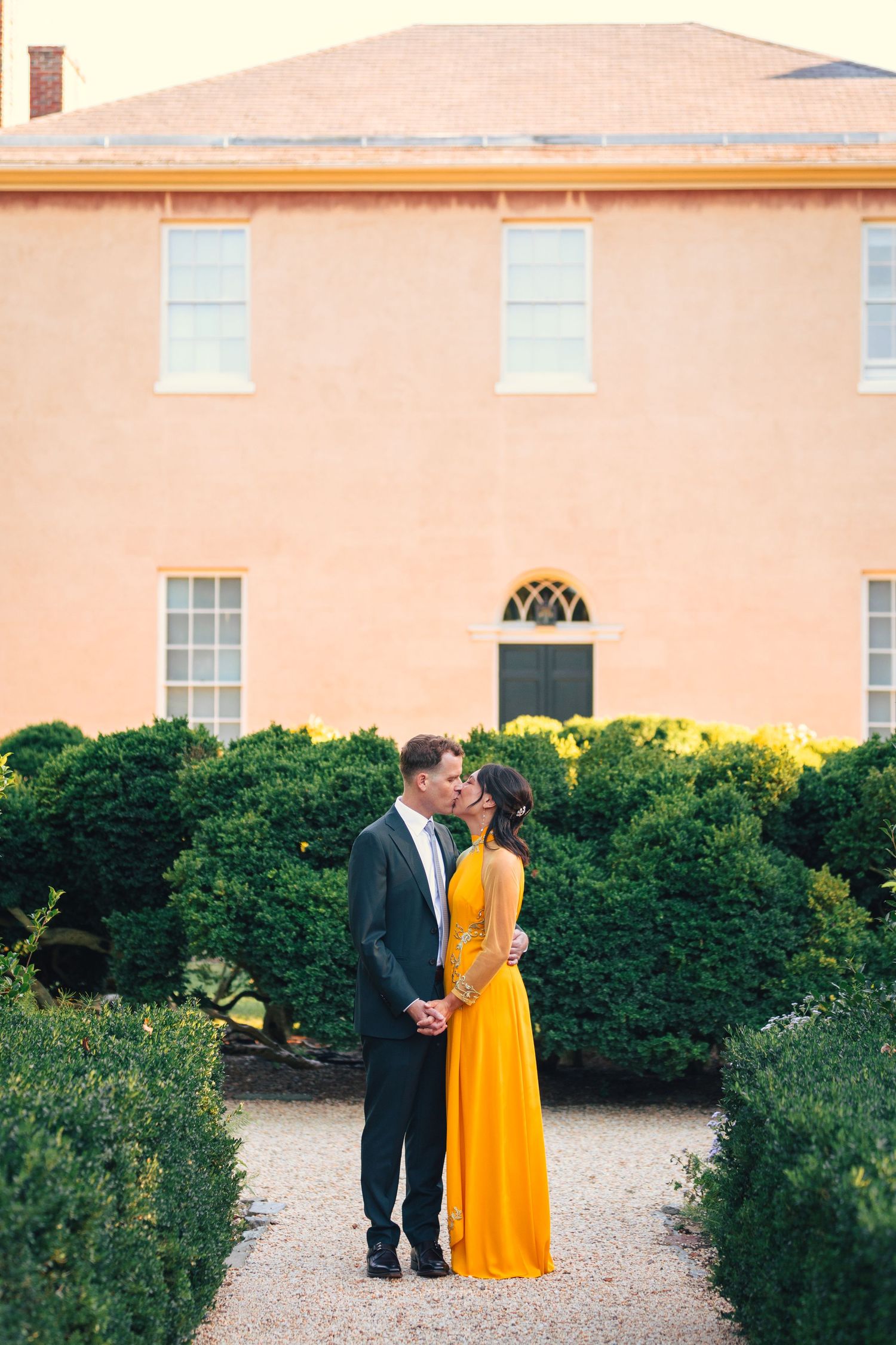Couple shares intimate moment in front of pink historic building with manicured gardens.
