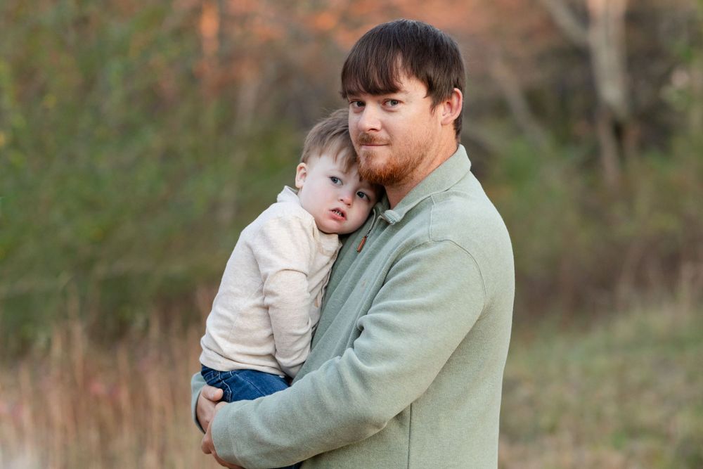 A tender moment captured outdoors as a parent holds a small child in a light blue sweater against an autumn background.