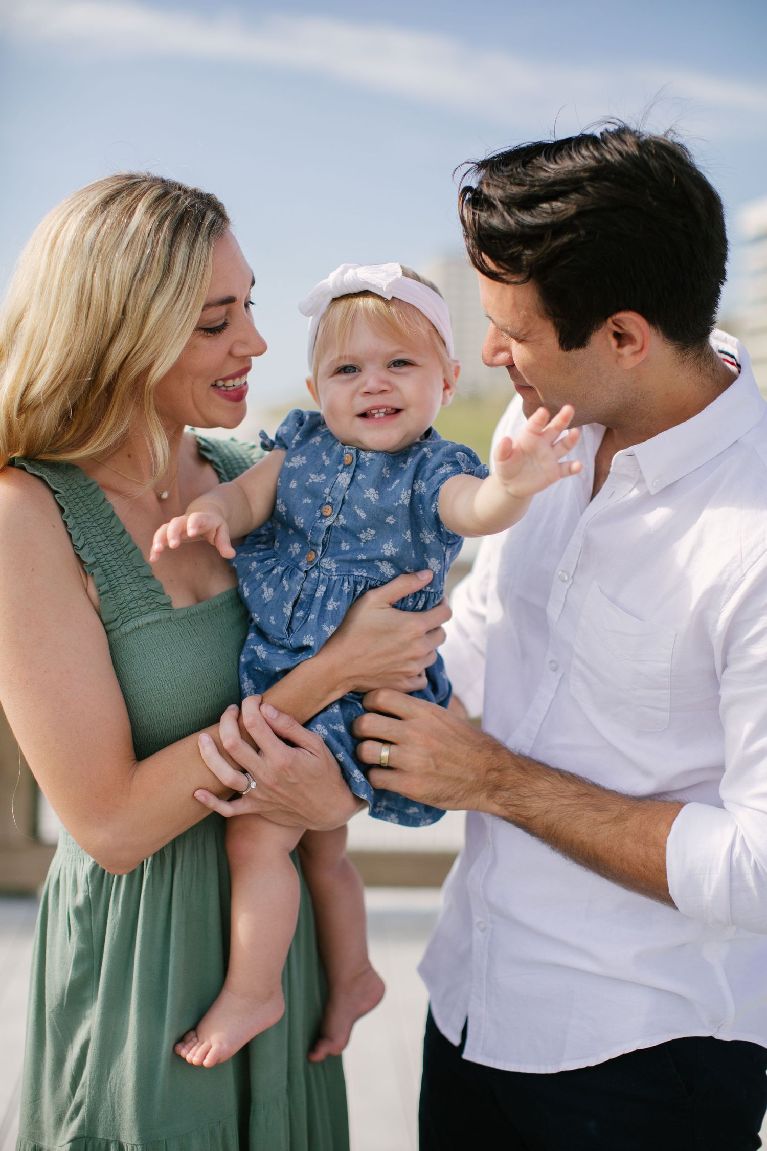 Happy family shares joyful moment outdoors with baby in blue dress and white bow headband.