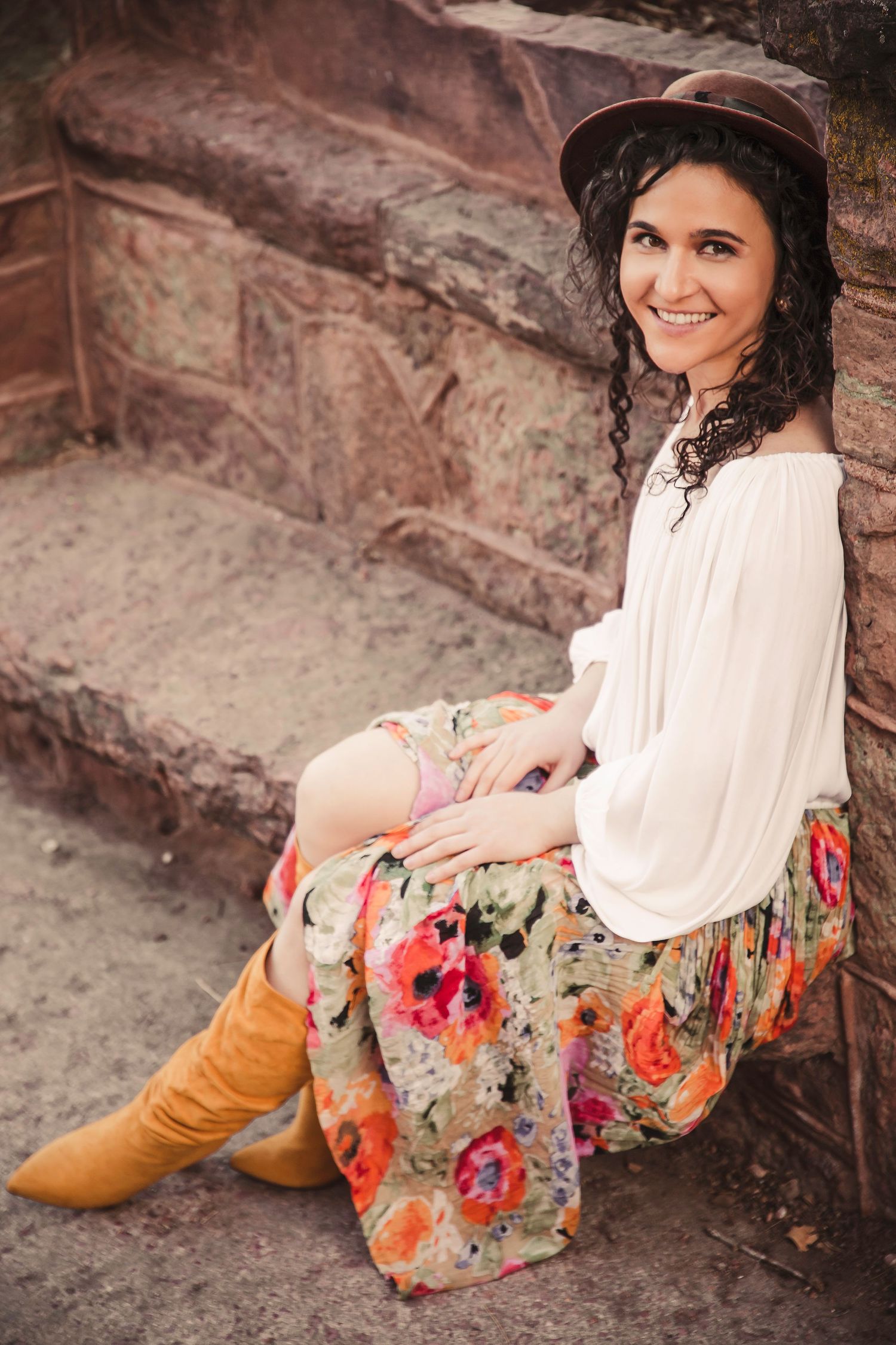 Woman in boho style dress and hat sits on red stone steps with floral pattern skirt.