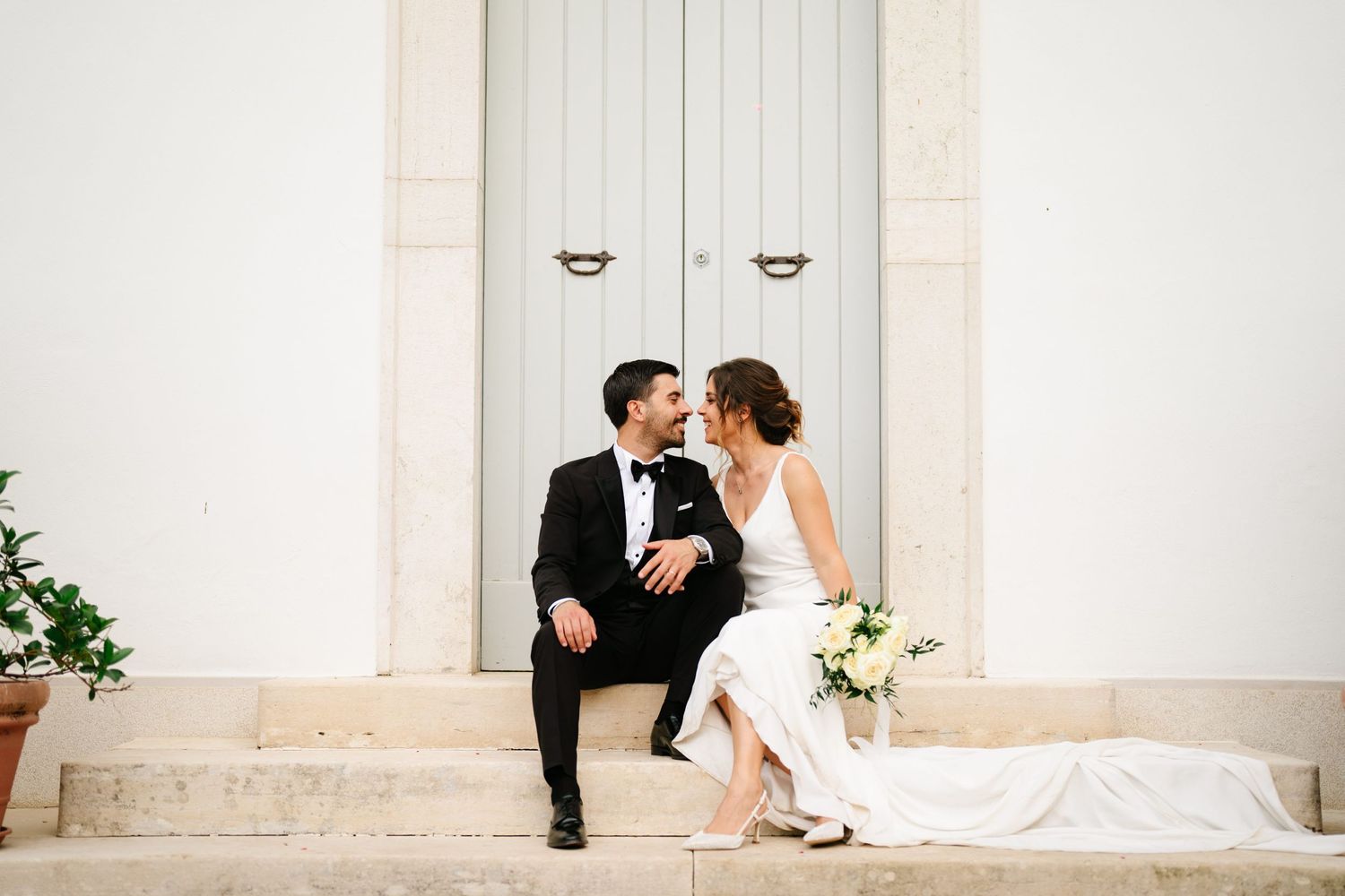 A playful wedding portrait of a couple sitting on white stone steps outside a classic building.