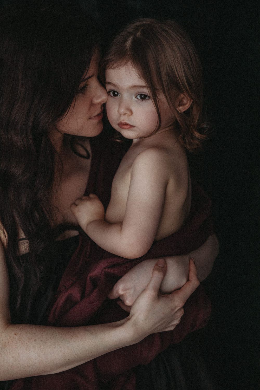 Woman holding a toddler in a close portrait against a dark background, photographed in soft light.