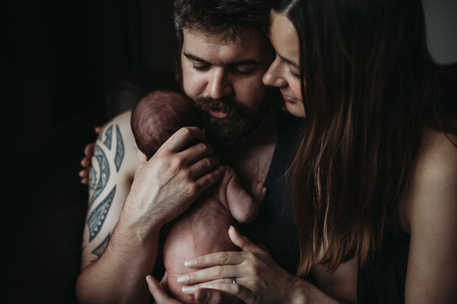 Parents holding their newborn baby in a close, intimate portrait photographed in low light.