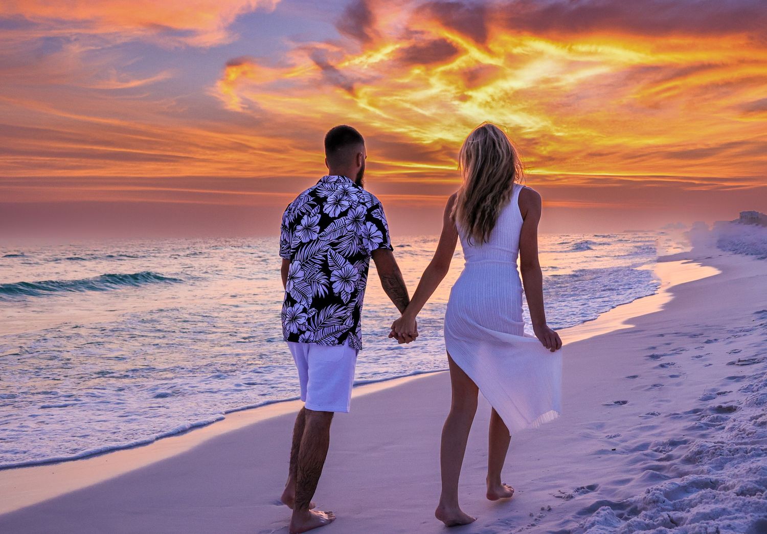 Couple walks hand-in-hand on white sandy beach against dramatic orange and purple sunset sky.