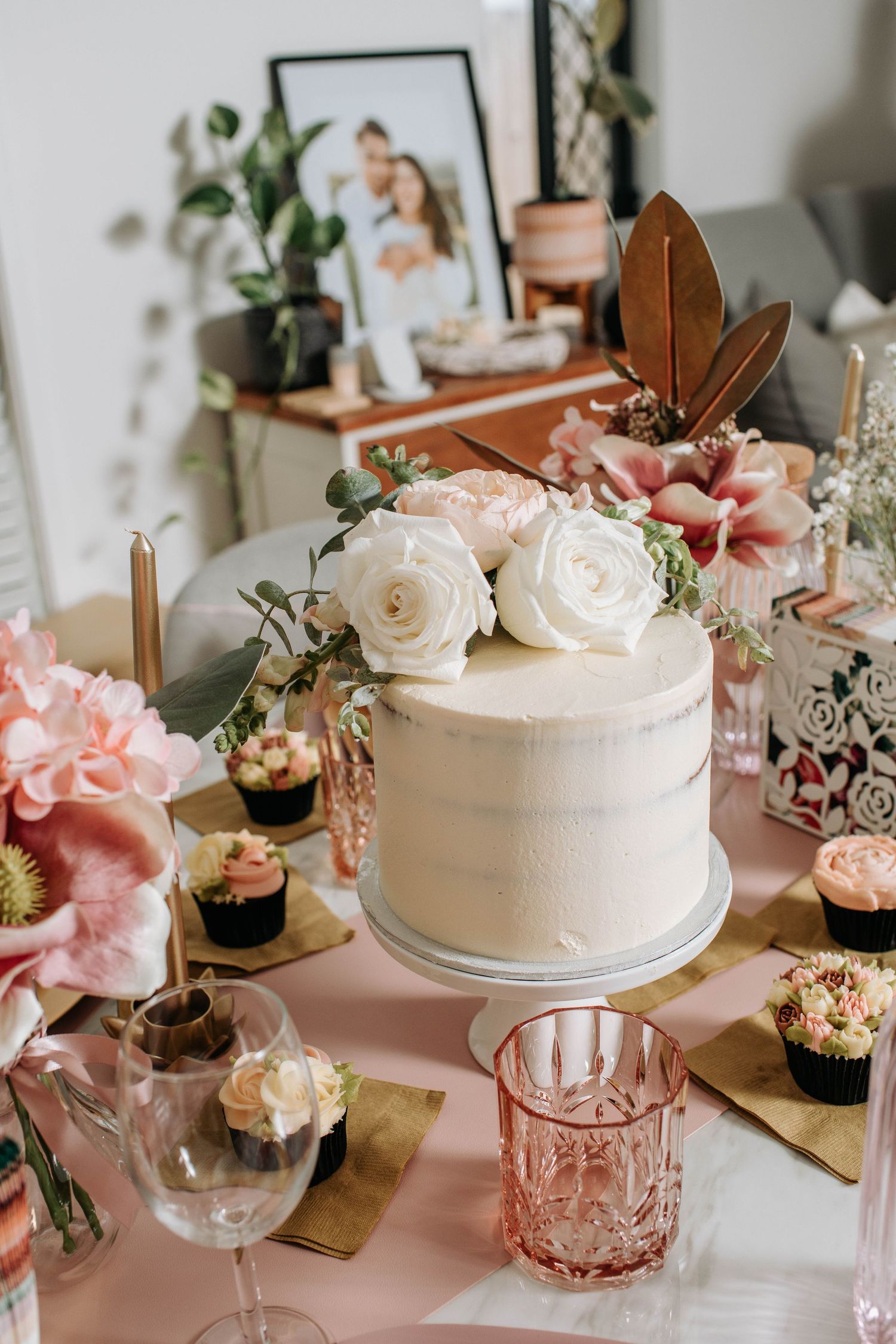 An elegant white wedding cake with roses and cupcakes on a pink tablescape.