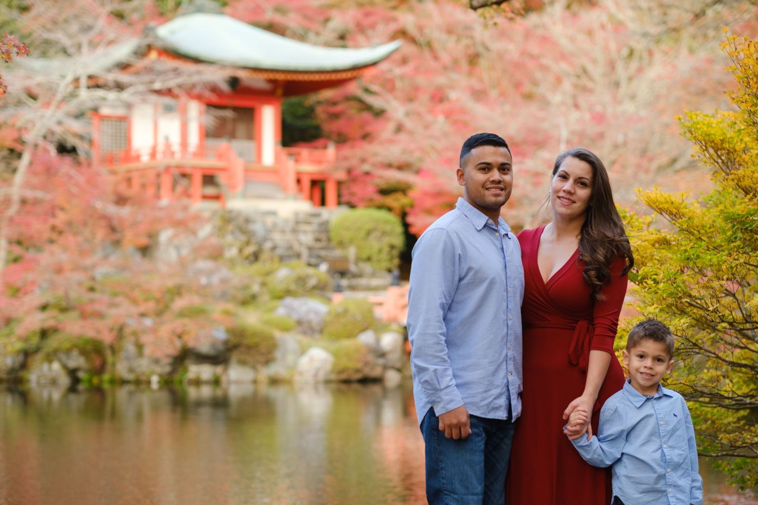 Family of three posing before a red Japanese pagoda reflected in a pond amid brilliant autumn foliage.
