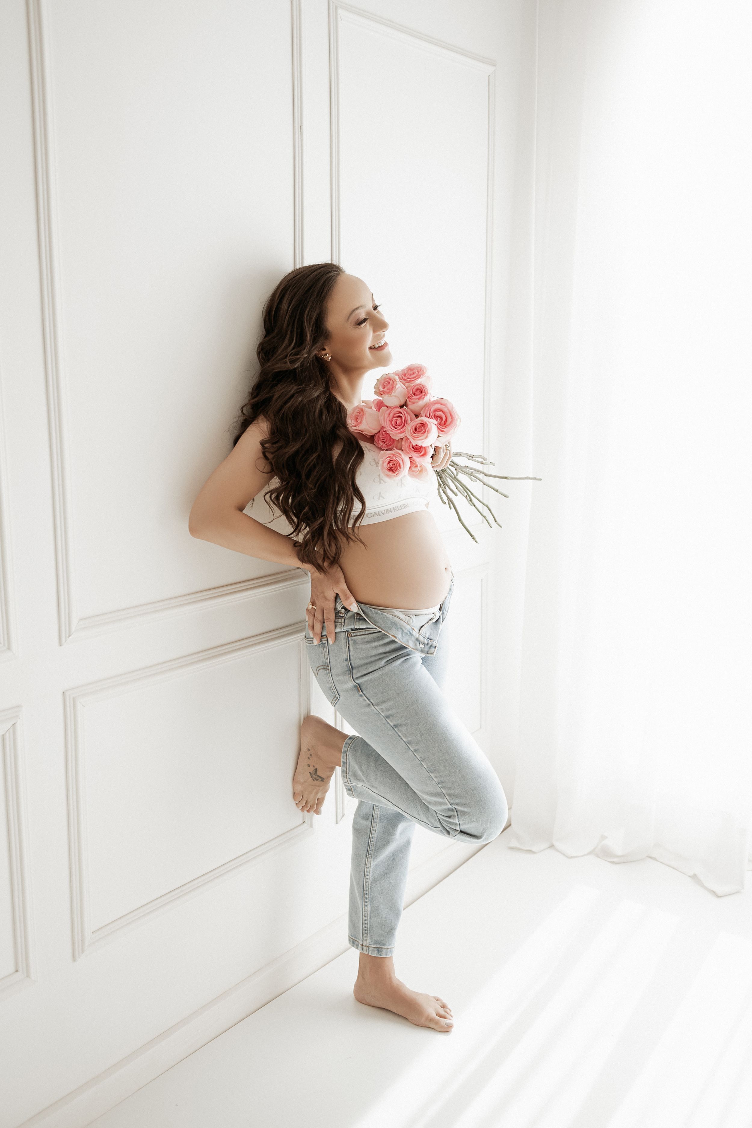 Expectant mother in casual denim holding pink flowers against white wall in natural light.