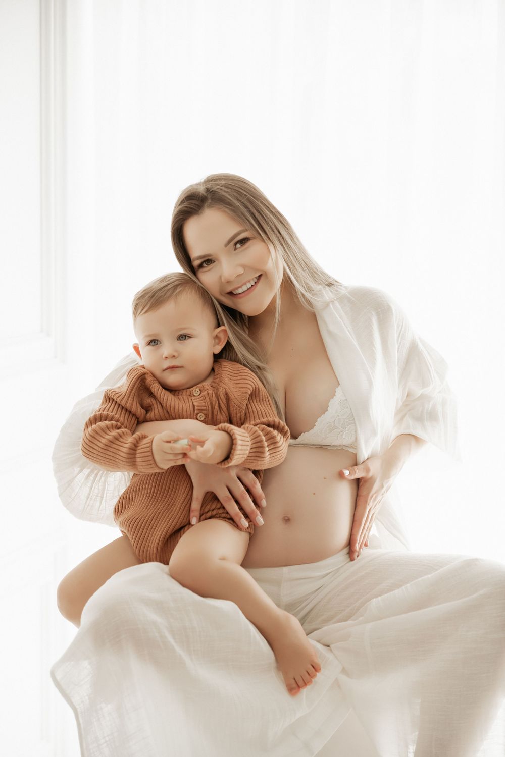 Intimate maternity portrait of parent and toddler in sweater against bright window.
