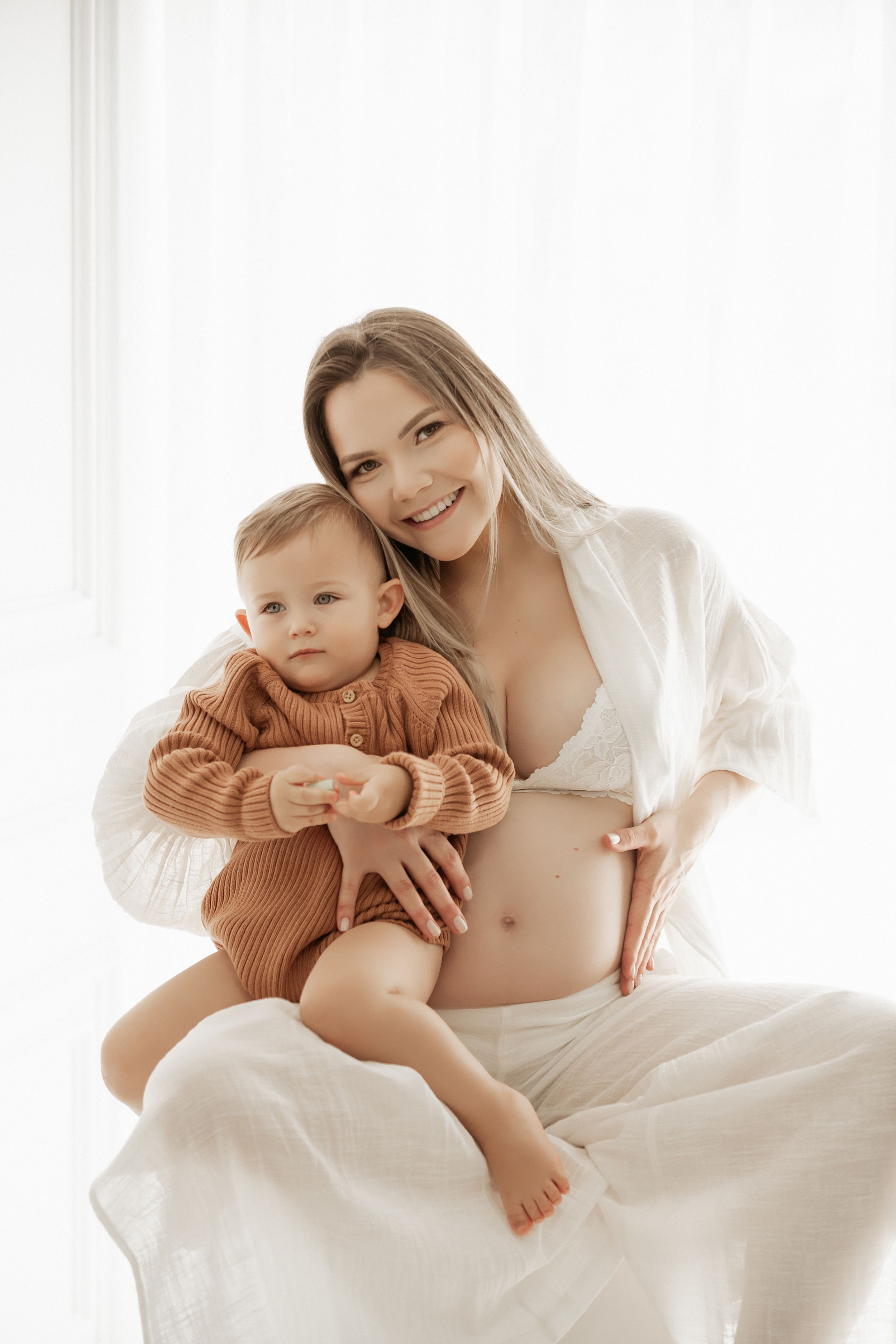 Intimate maternity portrait of parent and toddler in sweater against bright window.