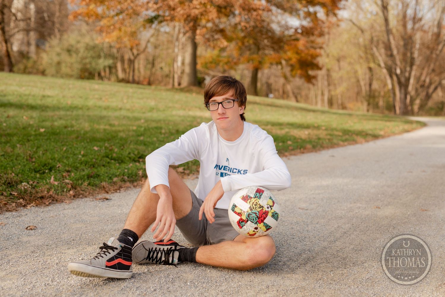 Student sits on path with decorative soccer ball in autumn park setting.
