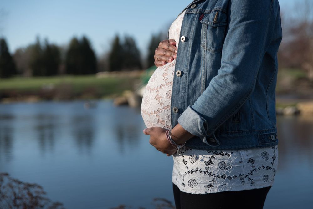 Close-up of pregnant woman in denim jacket and lace top holding belly near serene lake.