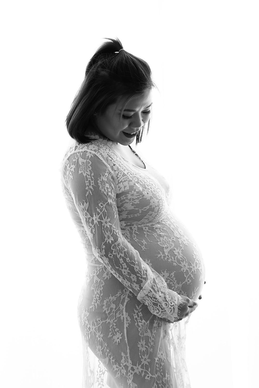 Black and white maternity photo of smiling pregnant woman in lace dress holding her belly.
