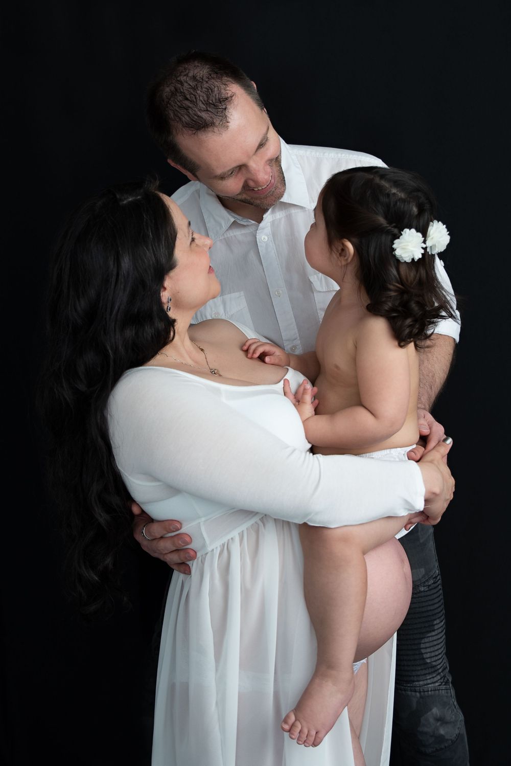 Family portrait of parents in white outfits lovingly holding their baby girl on black background.