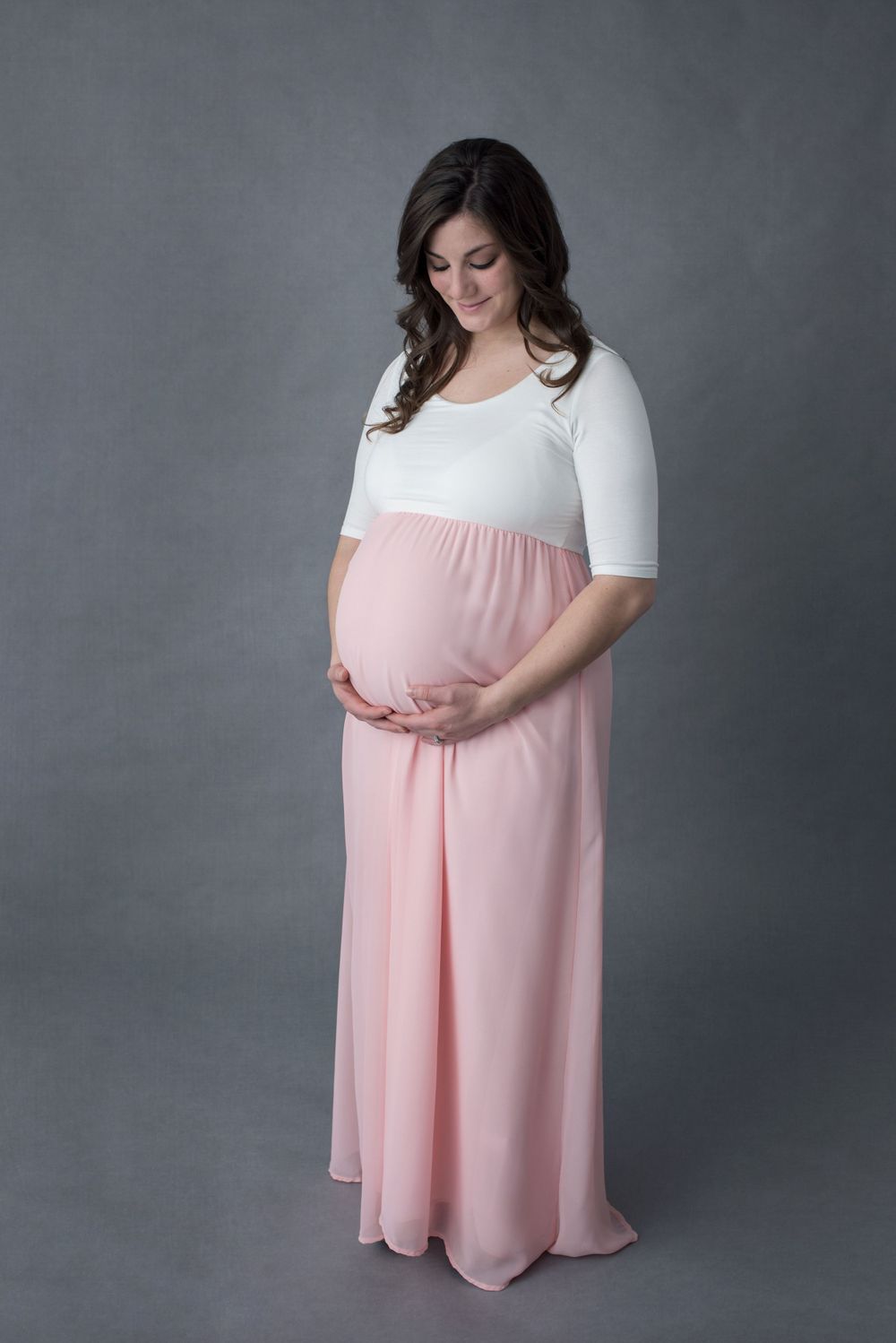 Pregnant woman in white and pink maternity gown cradling her baby bump in a studio.