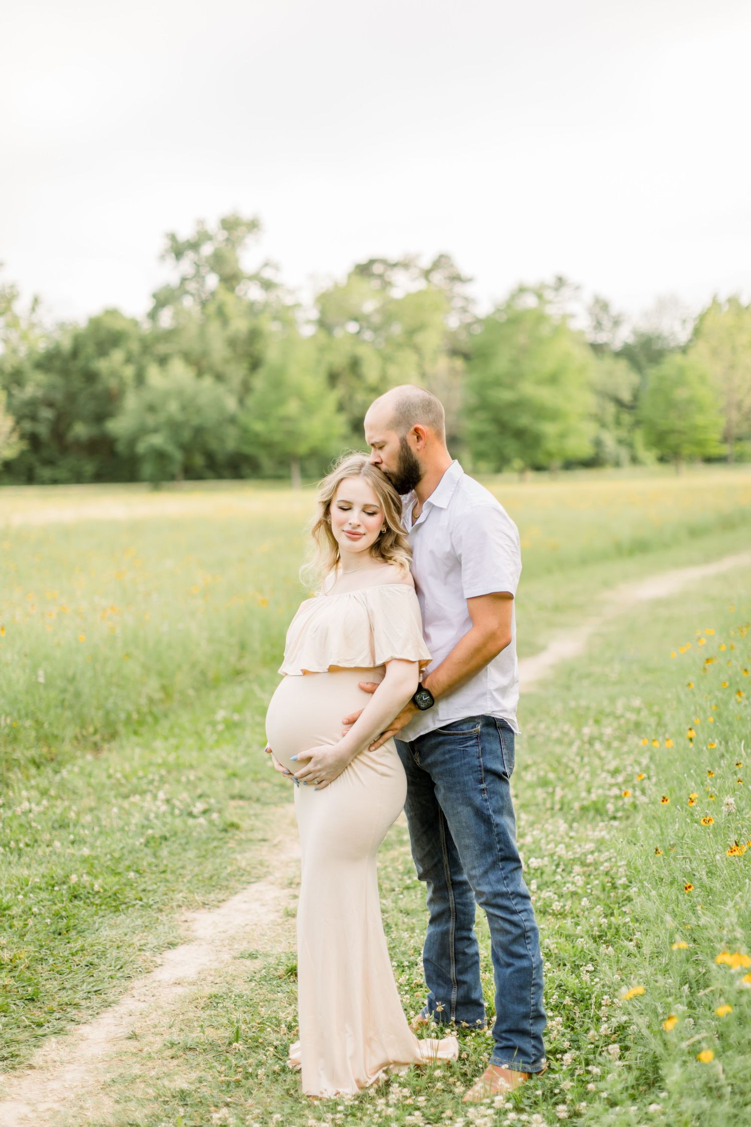 A couple shares an intimate moment during their maternity photoshoot on a dirt path surrounded by greenery.