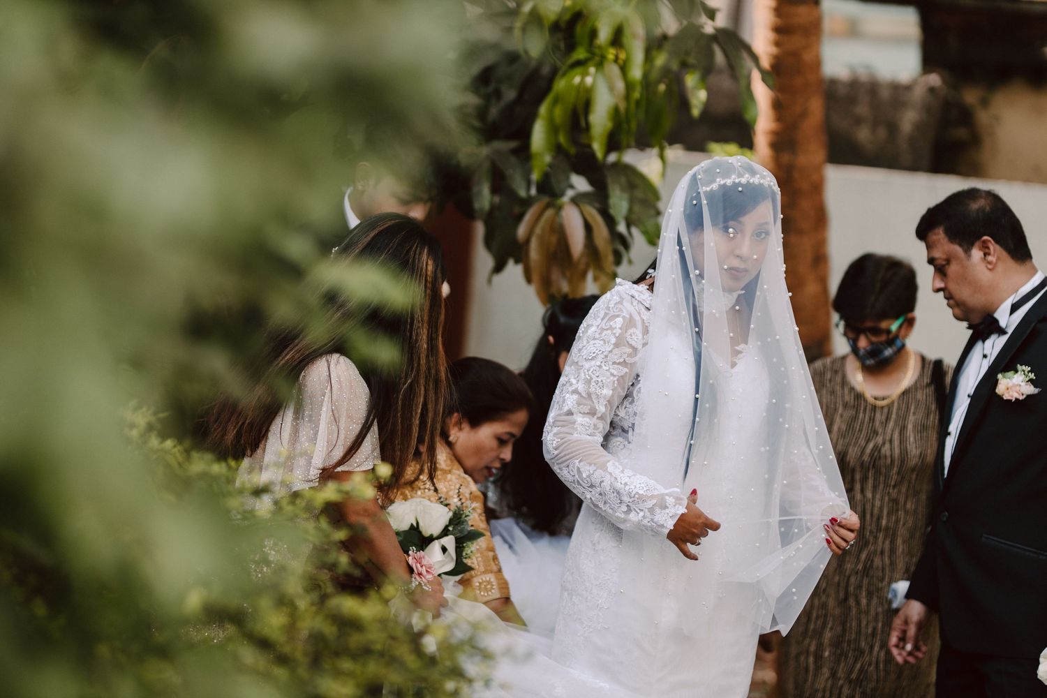 Wedding ceremony participants walking through an outdoor garden setting with natural foliage.