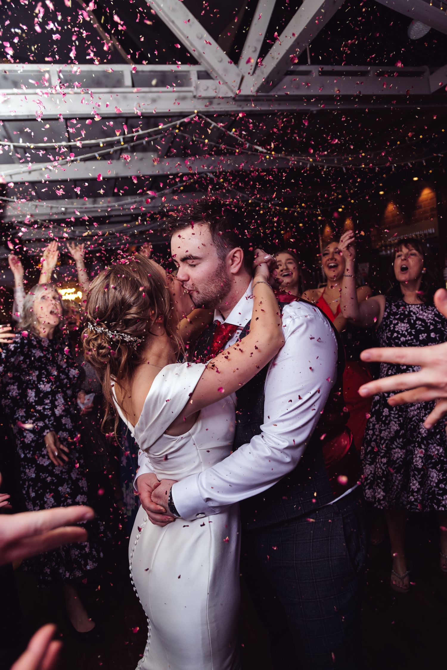 Newlyweds share a romantic kiss under falling pink confetti during their wedding celebration.