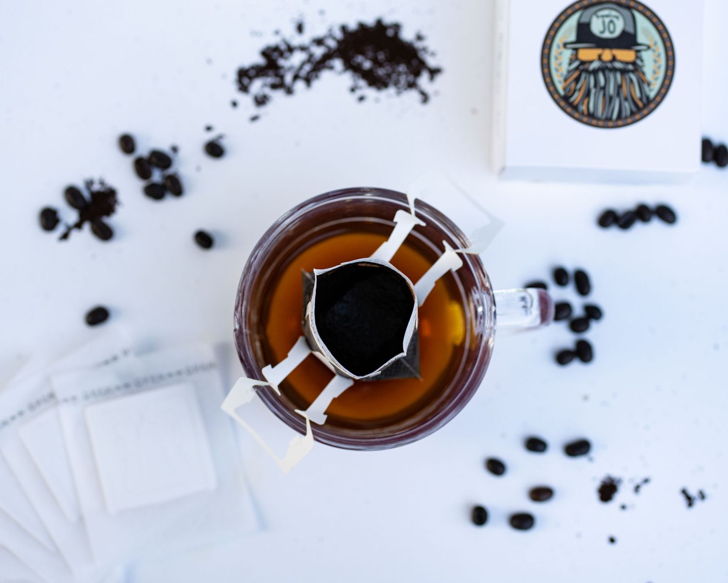 A box with logo, coffee beans, portable coffee filters, glass mug, and freshly poured coffee flat detail layout