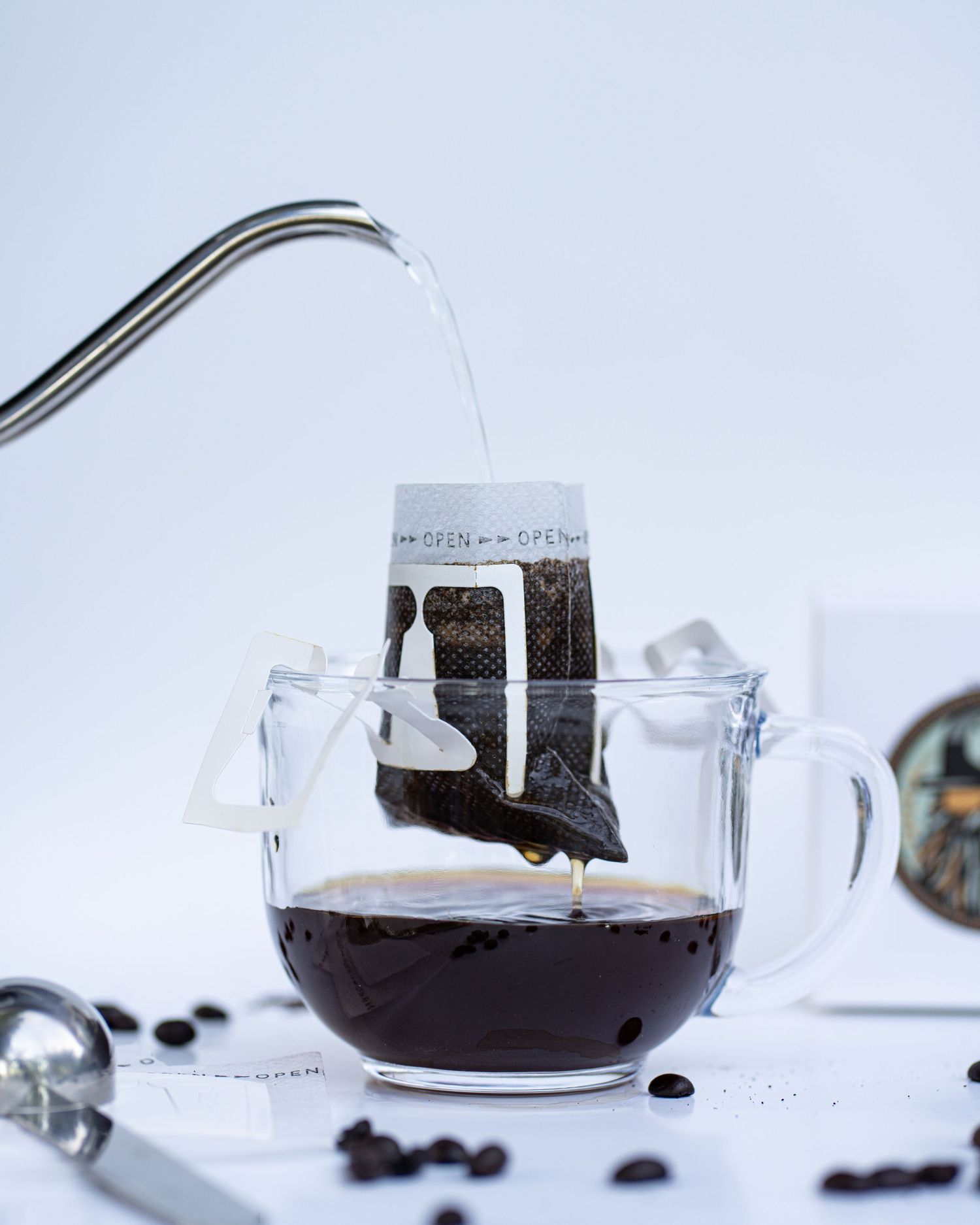 A kettle pours hot water over a portable coffee filter in a glass mug and coffee drips with box, beans, and scoop