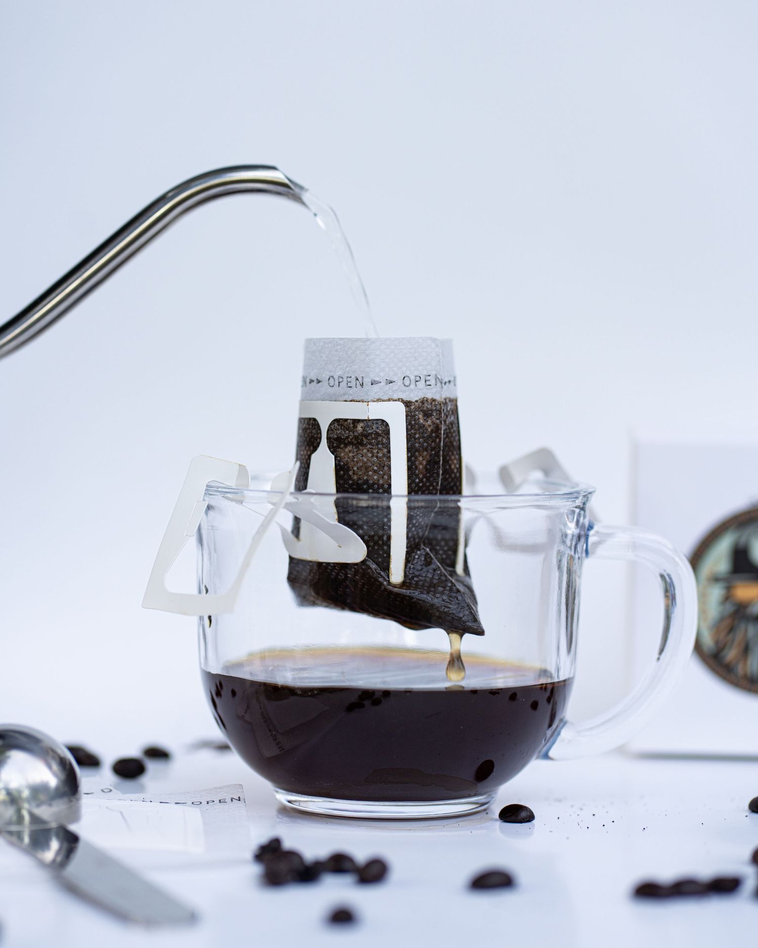 A kettle pours hot water over a portable coffee filter in a glass mug and coffee drips with box and logo in background