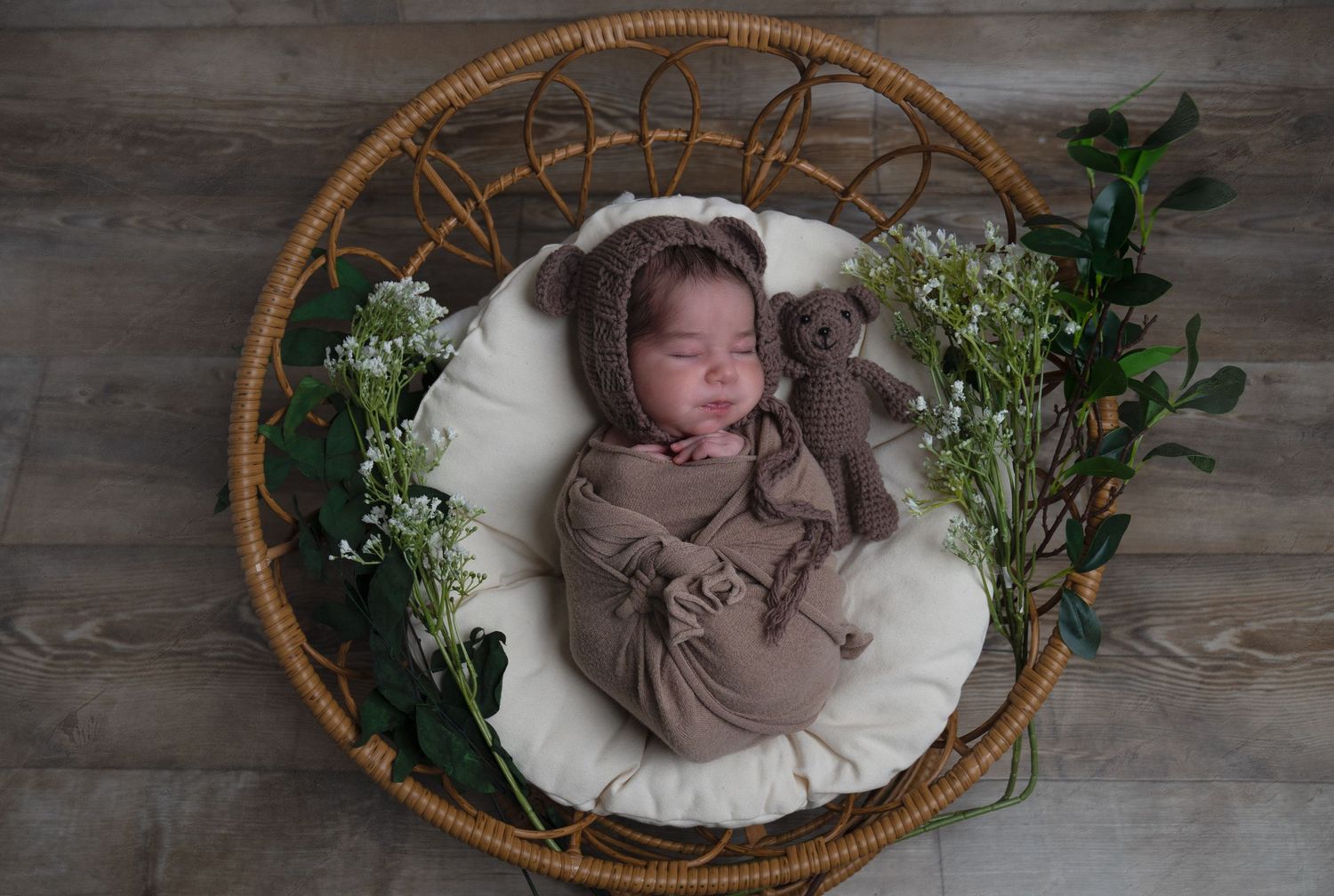 Sleeping newborn in brown wrap posed in wicker basket with greenery.