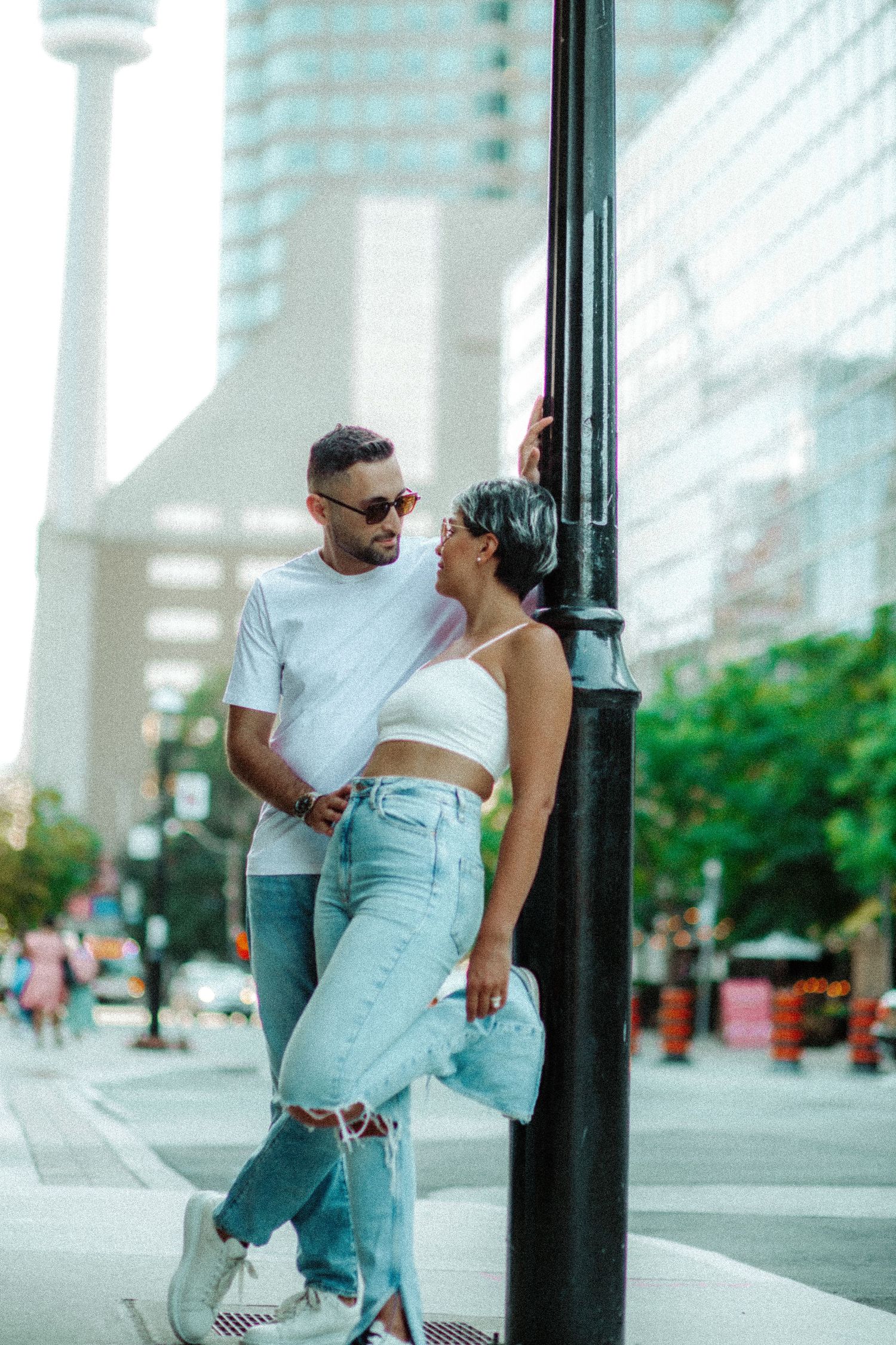A stylish couple poses against a lamppost in an urban setting with a city tower visible in the background.