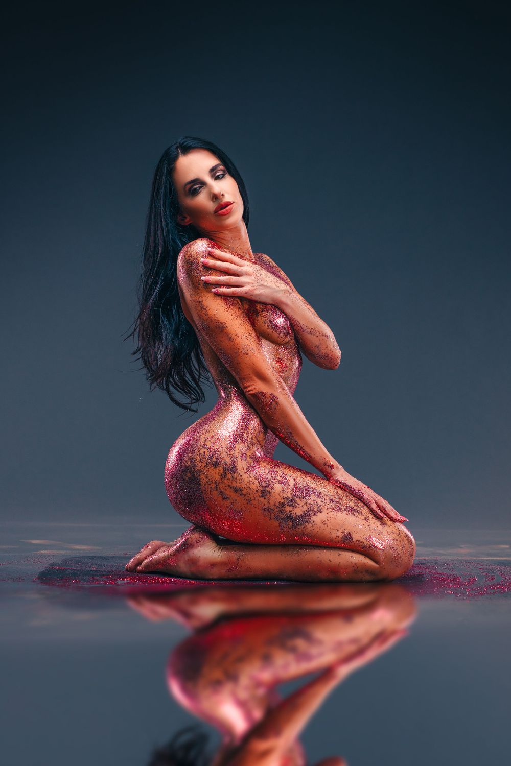 Artistic nude covered in glittering red body paint with mirror reflection on dark floor.