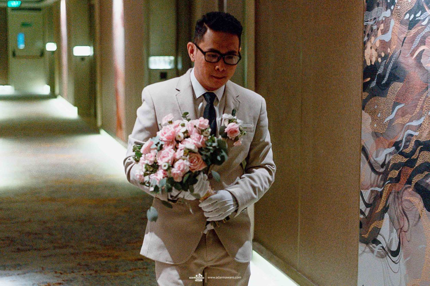 Figure in light colored suit carrying pink and white floral bouquet in hotel hallway.