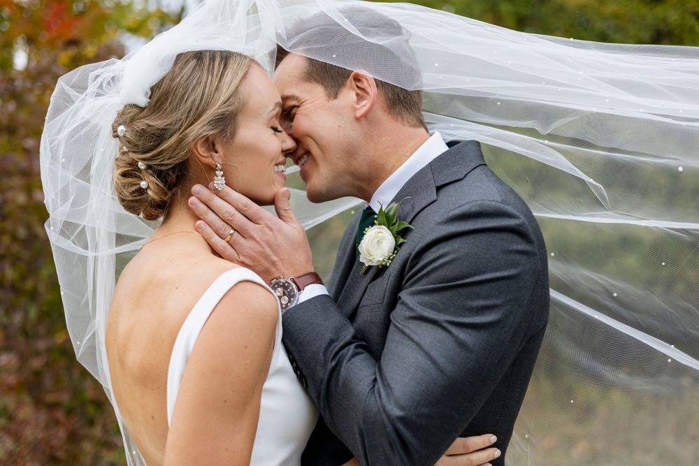 A romantic wedding moment captured outdoors with a flowing veil and fall foliage in the background.