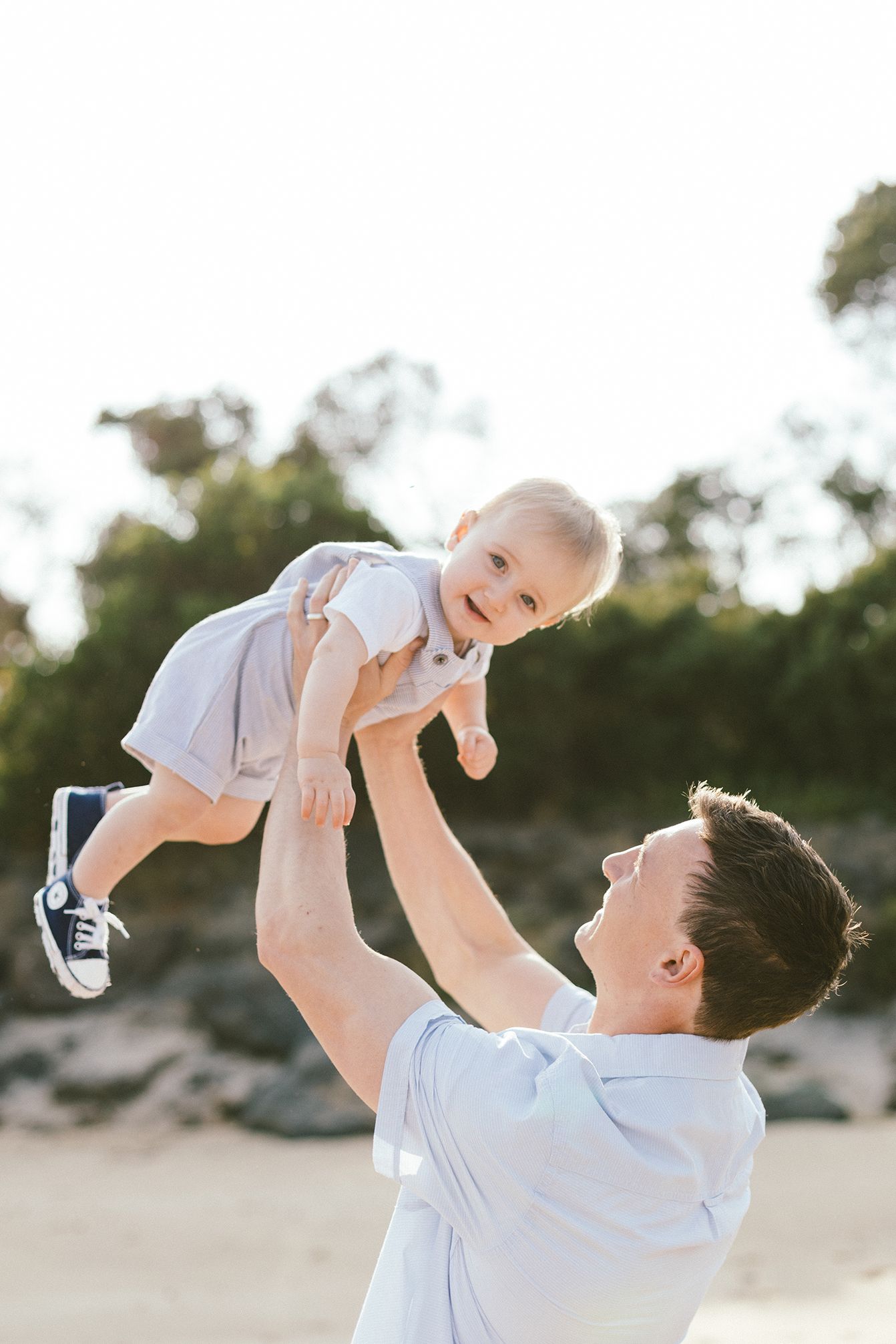 Father playfully lifts baby in white outfit against scenic beach background.