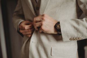 Detail shot of hands buttoning a beige suit jacket with watch visible.