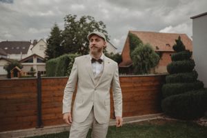 Beige-suited figure stands confidently in garden setting with brick wall and topiary.