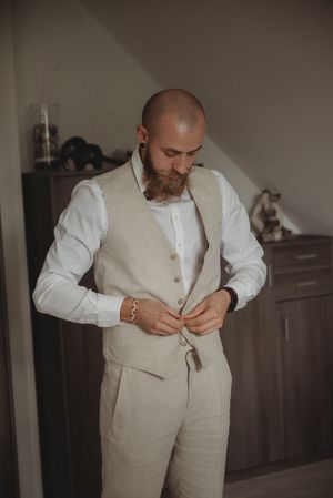 Person in beige suit and white dress shirt buttoning up waistcoat indoors.